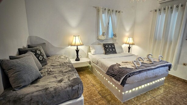 A bedroom with a bed, a sofa, two bedside tables with lamps, a window with curtains, and a door with a sign. - Pleasure Beachside Studios (Santorini)