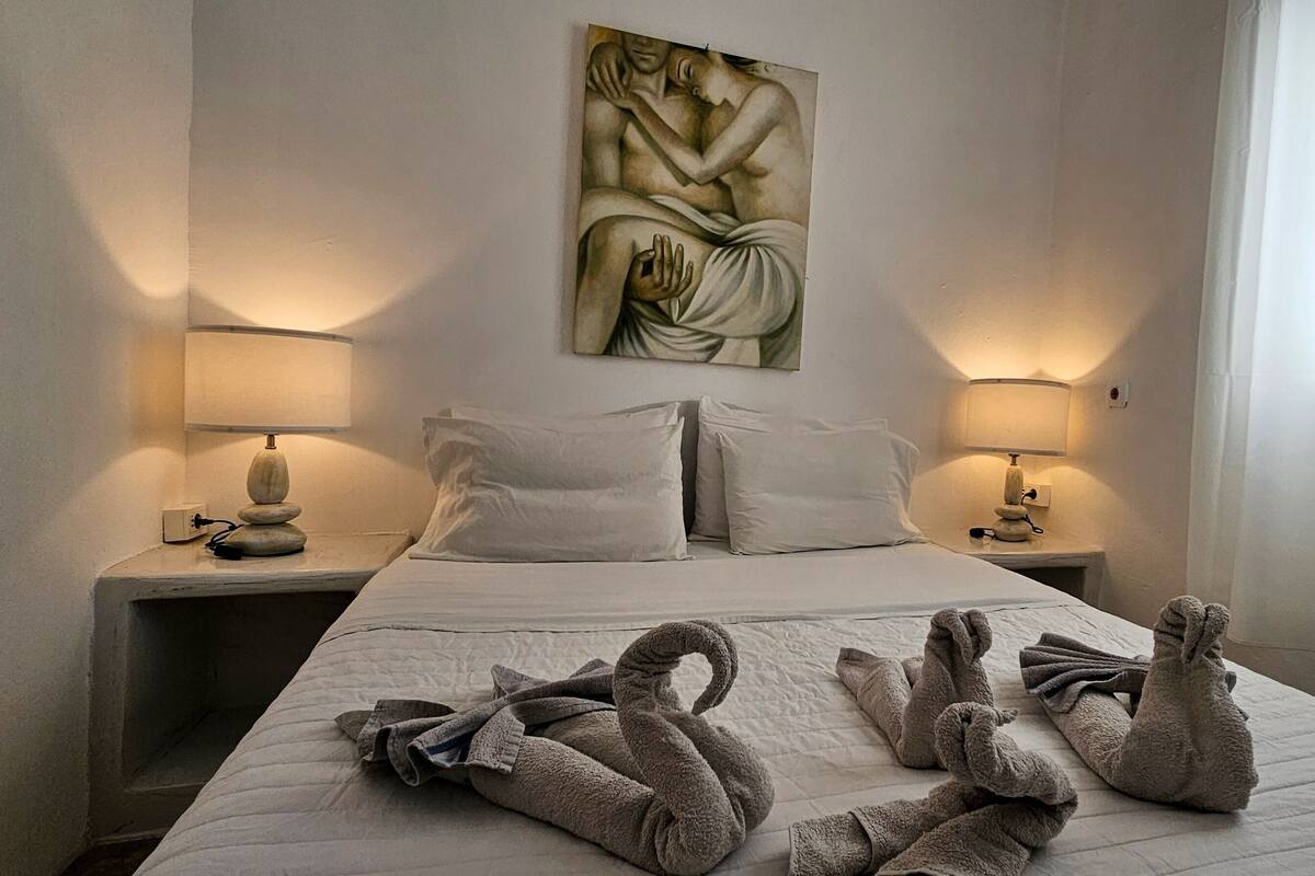 A hotel room with a bed, two bedside lamps, a painting on the wall, and towels arranged on the bed.