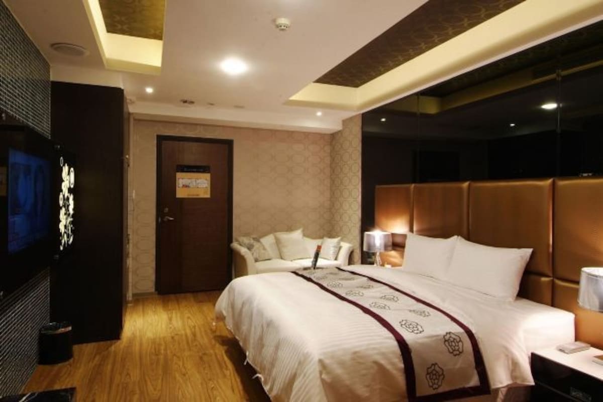A hotel room with a large bed, a TV, a sofa, and a desk with a lamp.