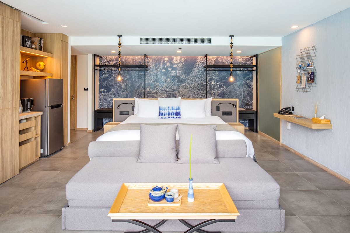 Une chambre d’hôtel moderne dotée d’un grand lit, d’une table de chevet en bois, d’un coin cuisine avec un réfrigérateur et offrant une vue sur la ville par les fenêtres.