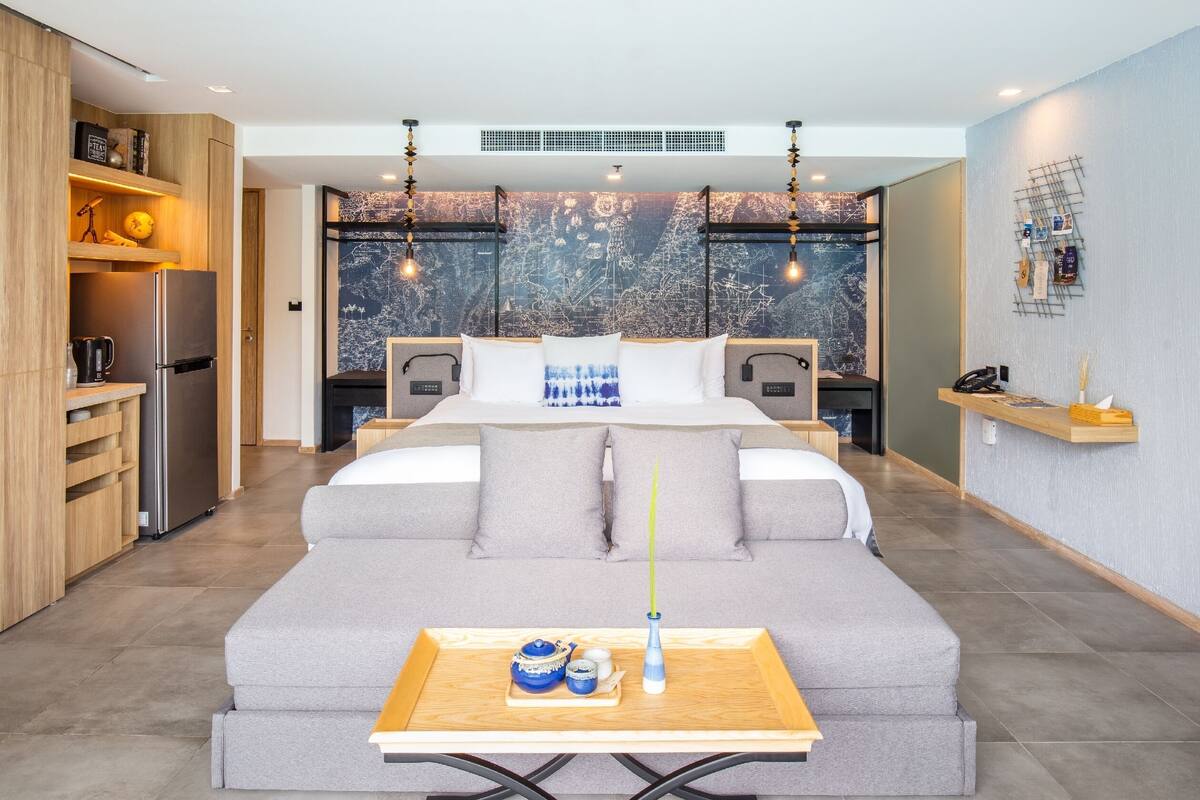 Een moderne slaapkamer met een slaapbank, een nachtkastje met een vaas en theepot, een groot raam met uitzicht op de stad, en een keuken met een koelkast en planken.