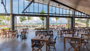 A spacious outdoor dining area with wooden tables and chairs, large windows offering views of the beach and palm trees.