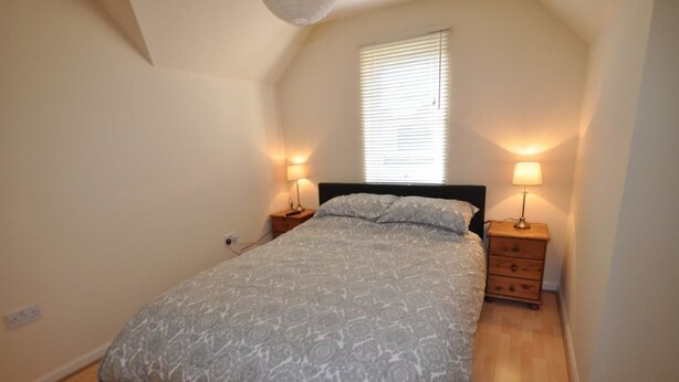 A bedroom with a bed, two bedside tables with lamps, a window with blinds, and a sloped ceiling. - 6 Varis Apartments (Forres)