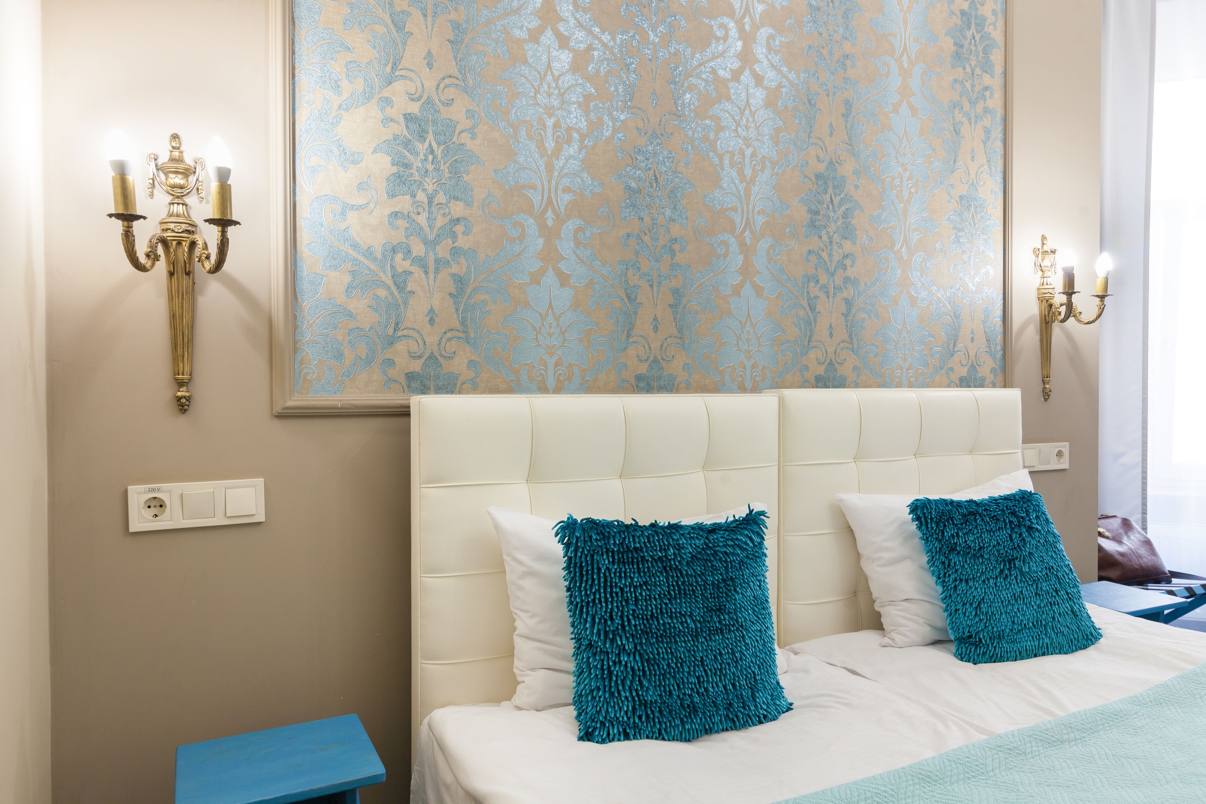 A hotel room with a bed, two teal pillows, a cream-colored headboard, a blue nightstand, and a wall with a patterned wallpaper.