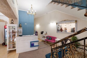 Een ruime woonkamer met een trap, een kroonluchter, een blauwe accentmuur, een roze bank en een witte kast met glazen deuren.