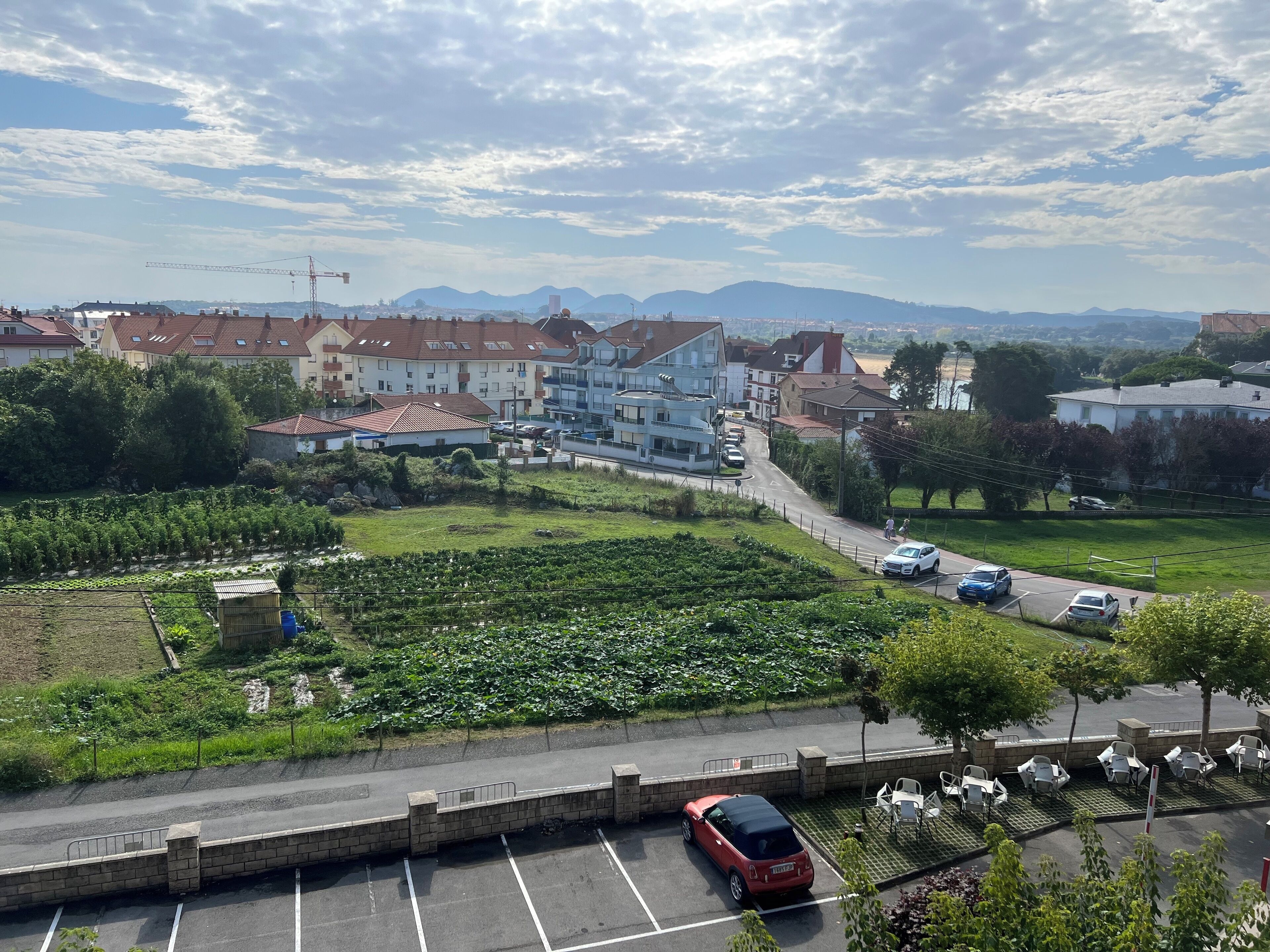 Un estacionamiento con un coche rojo, un coche blanco y un coche azul. Se ven edificios al fondo y una vista de montañas.