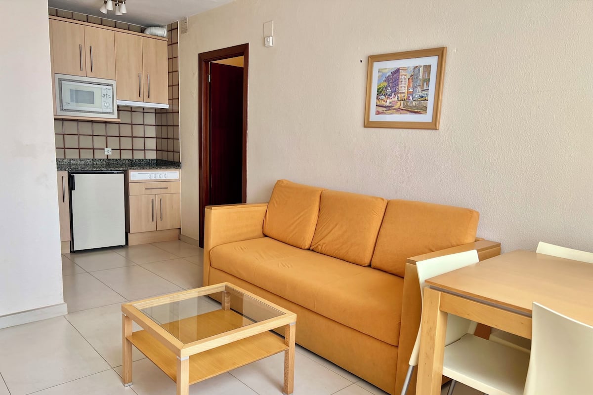 A compact living space with a sofa, a glass-top coffee table, a dining table with chairs, a microwave, and a refrigerator.