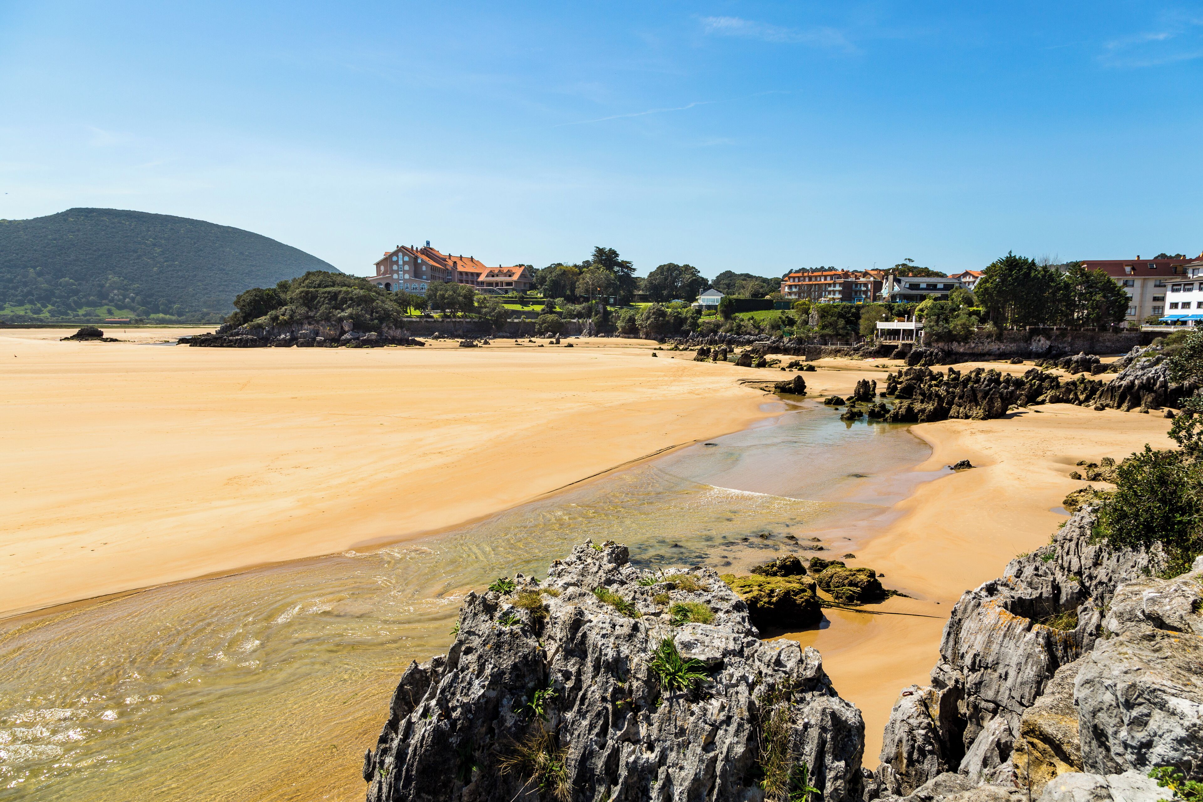 A coastal beach with rocky outcrops and buildings in the background.