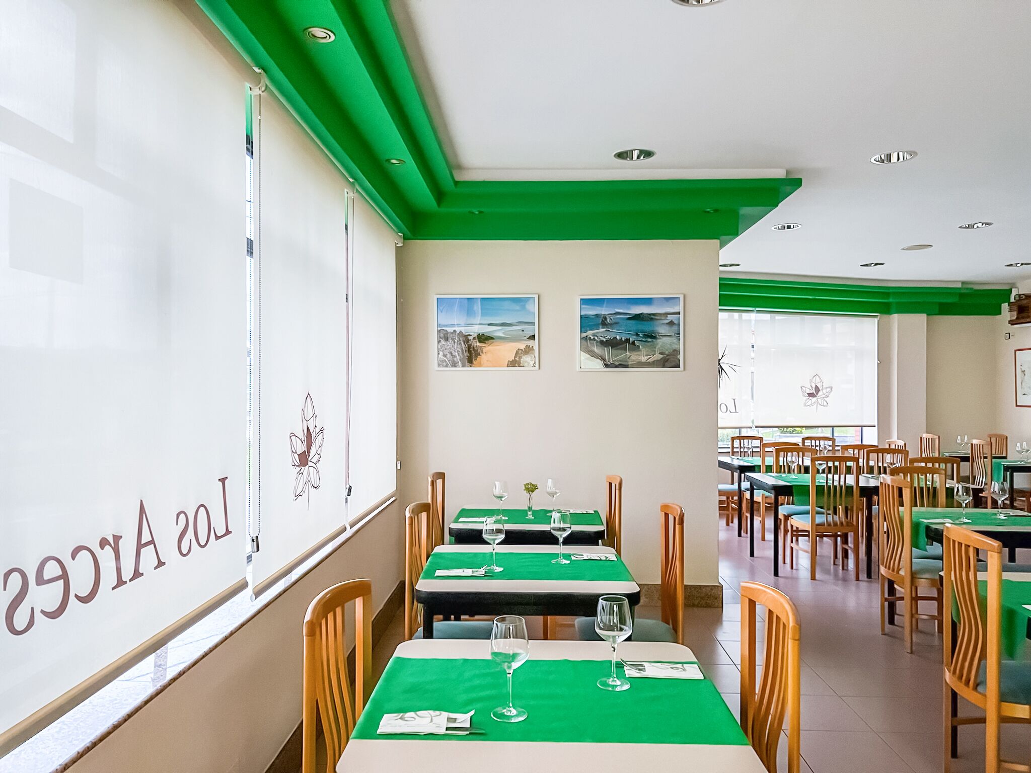Un restaurante con manteles verdes, sillas de madera y cuadros de paisajes costeros en las paredes.