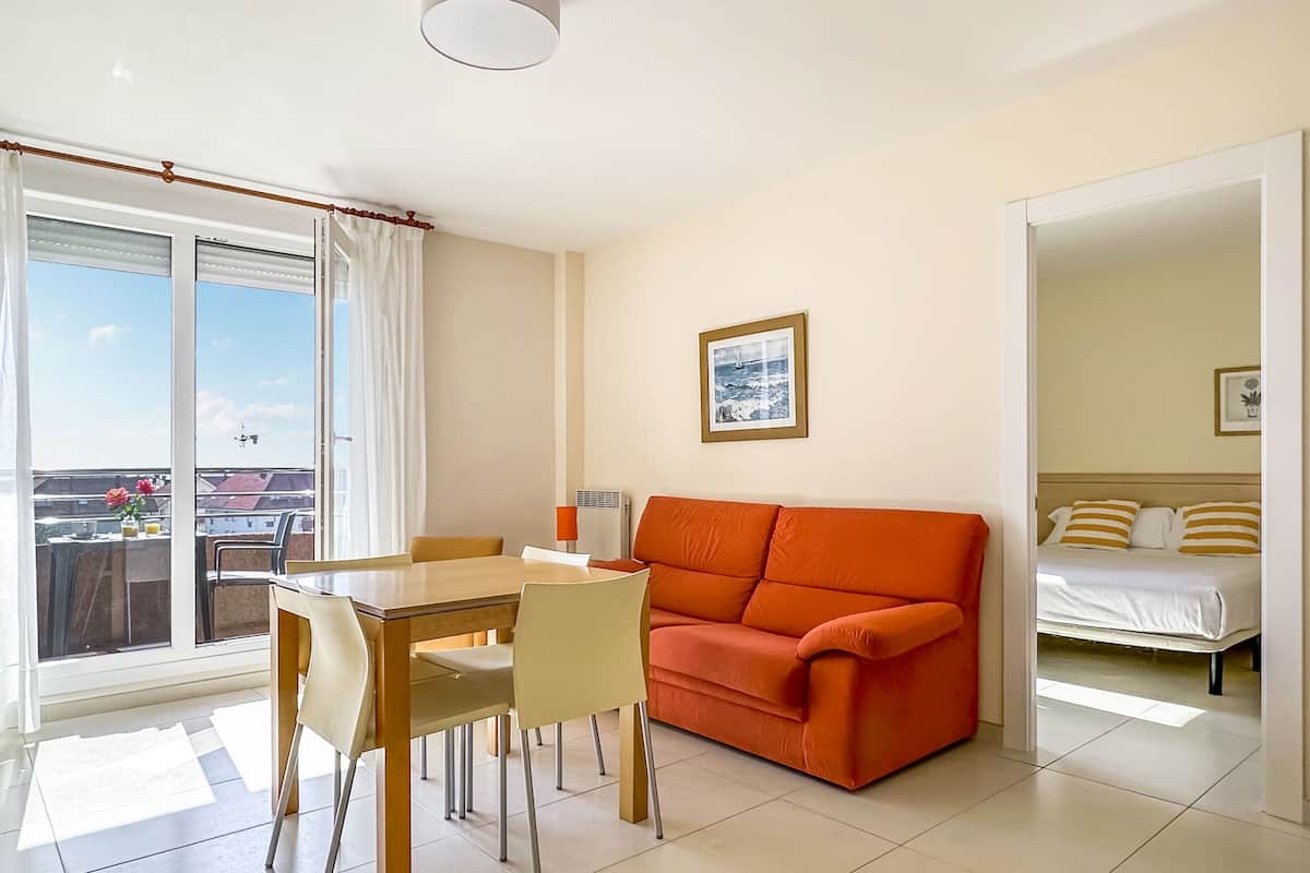 A room with a dining table, chairs, an orange sofa, a bed, and a window with a view of buildings and a clear sky.