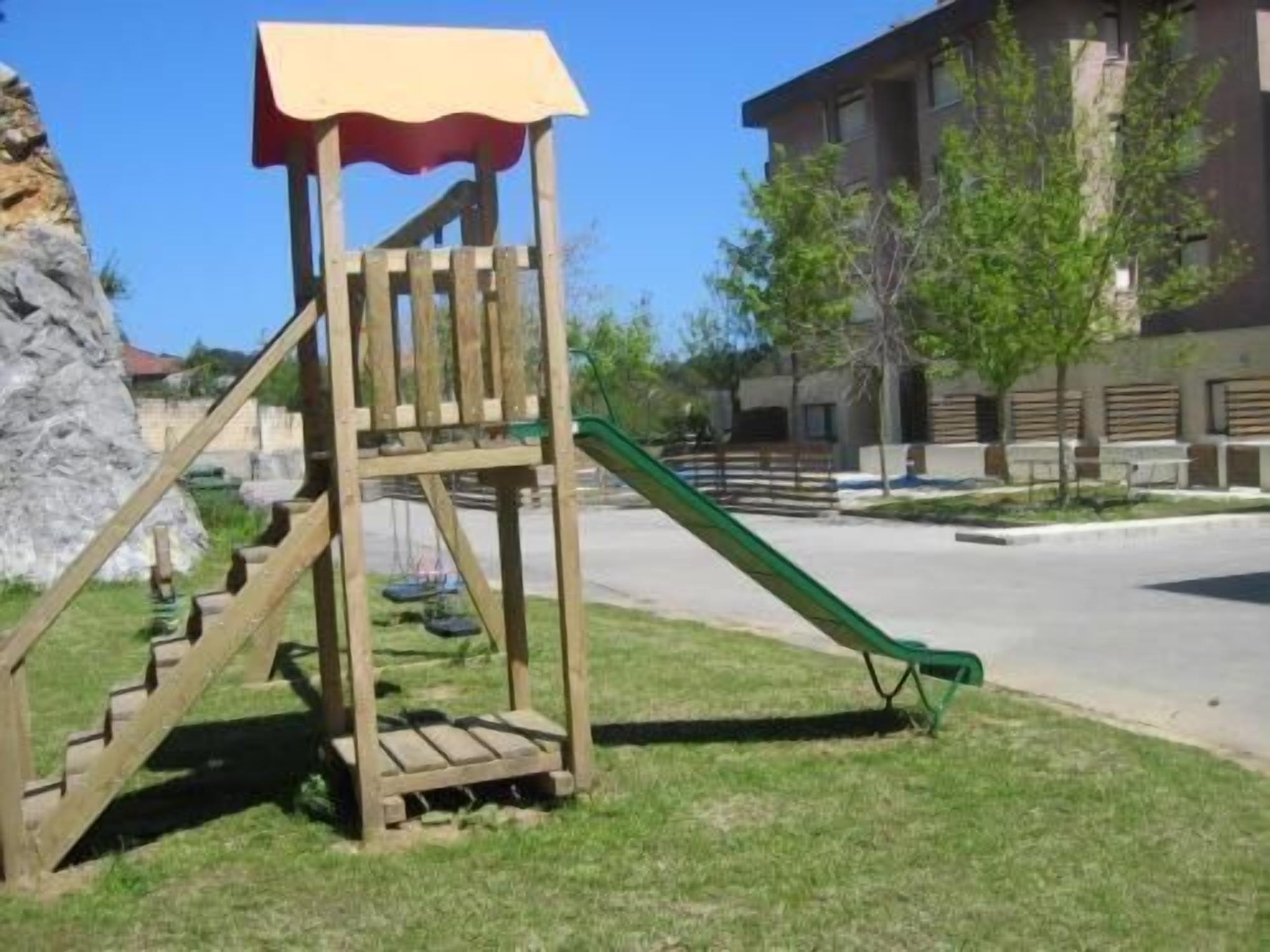 Una struttura giochi in legno con uno scivolo, immersa in un prato con un edificio sullo sfondo.