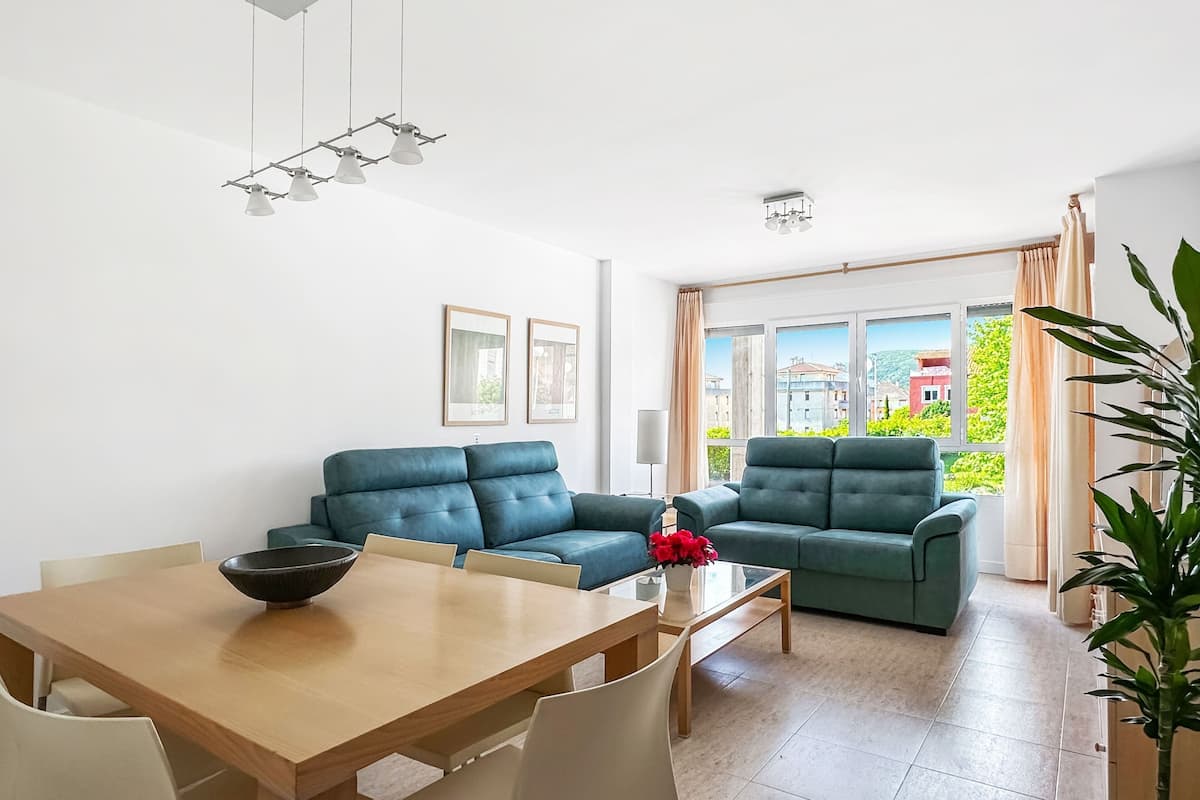 Una sala de estar moderna con una mesa de comedor de madera, dos sofás y un amplio ventanal con vistas a edificios y vegetación.