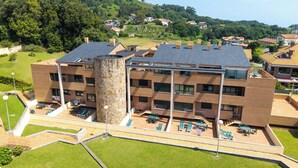 A multi-story building with a central tower, outdoor seating area, and a view of a hillside with houses. - Apartamentos Solamaza (Arnuero)