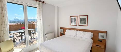 A hotel room with a bed, a chair, a table with flowers, a radiator, and a view of the city through a large window.