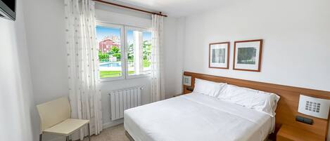 A hotel room with a large bed, a chair, a bedside table, a window with curtains, and two framed pictures on the wall.