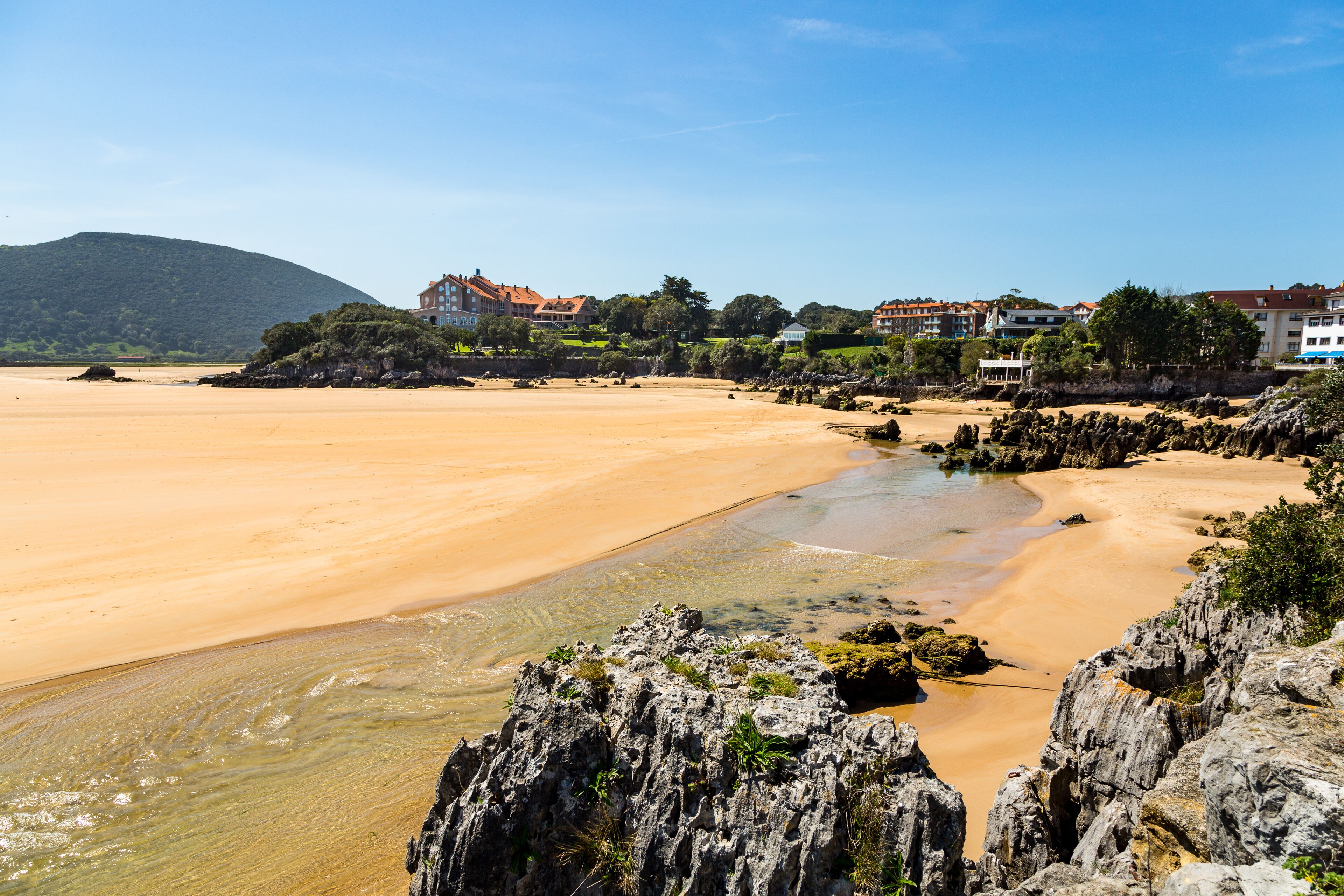 A coastal beach with rocky outcrops and buildings in the background.