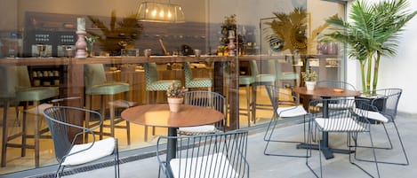 An outdoor seating area with round tables and metal chairs, a potted plant, and a view of the interior through a glass partition.