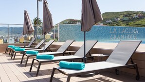 Un área de lounge en la azotea con sillones, toallas y sombrillas, con vistas panorámicas y un cartel de hotel al fondo.