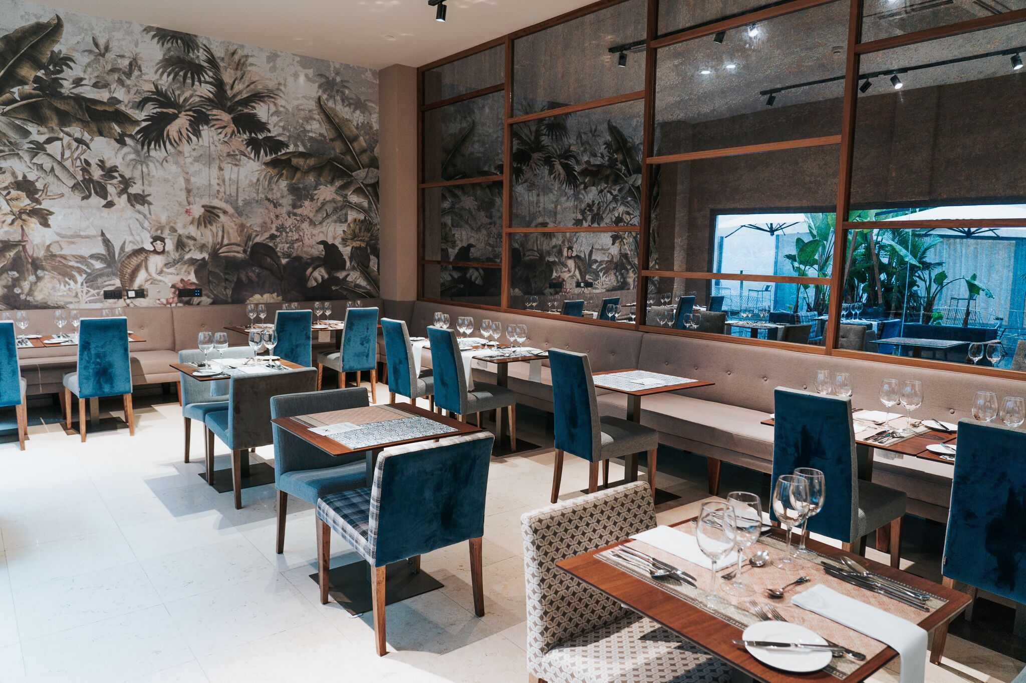 Un restaurant décoré d’une fresque aux motifs tropicaux, doté de chaises en velours bleu et de tables en bois.