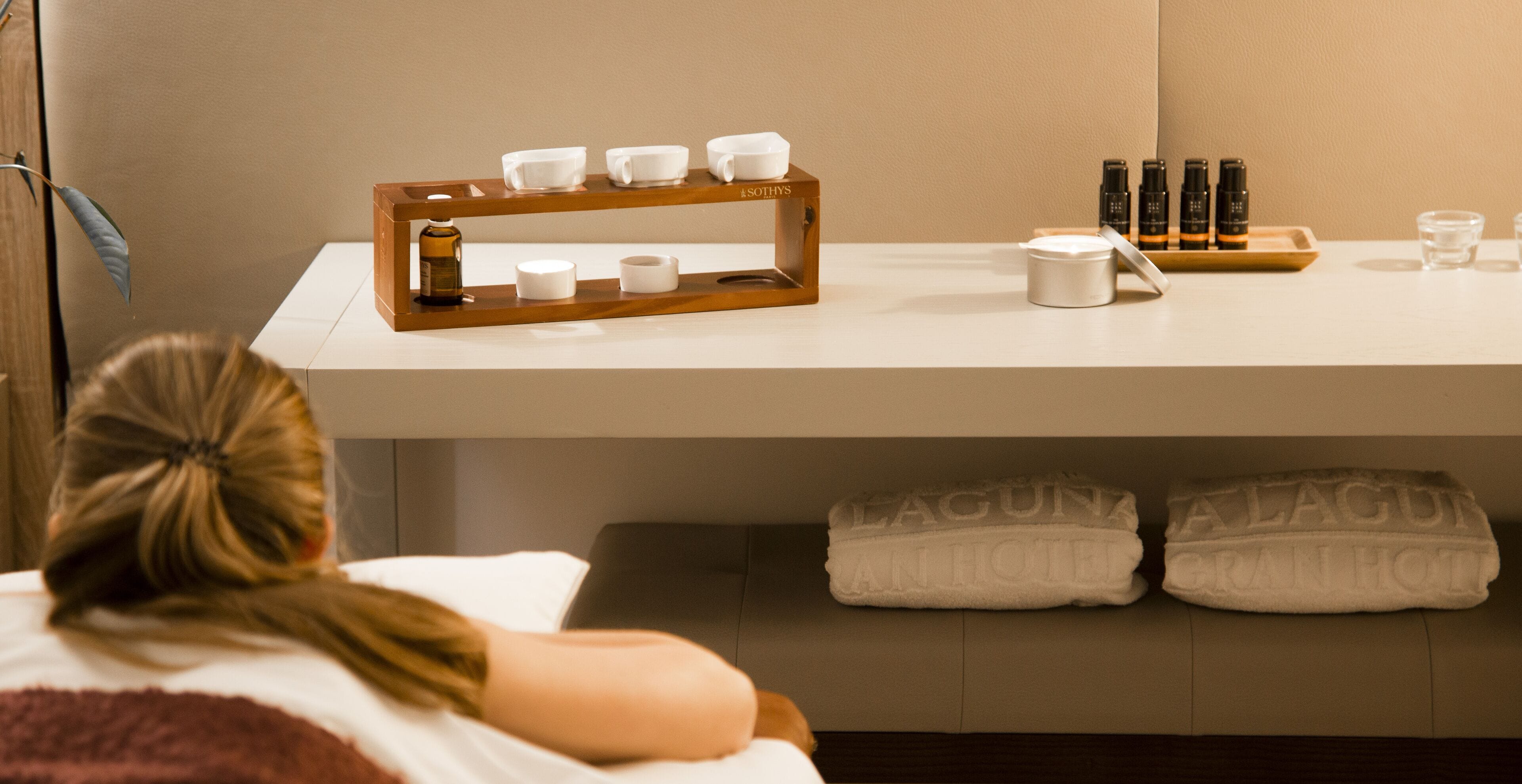 Ein Hotelzimmer mit einem Holftablett, das weiße Tassen, eine Flasche, einen kleinen weißen Behälter und ein Glas auf dem Tisch zeigt. Auf dem Bett liegen zwei Handtücher mit dem Schriftzug "Laguna".