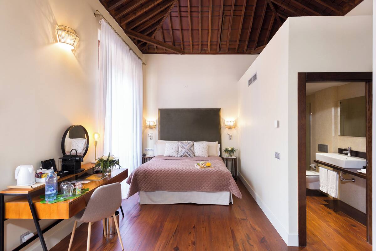 A bedroom with a bed, bedside tables, a desk, a chair, a mirror, a window with curtains, and a bathroom with a sink and mirror.