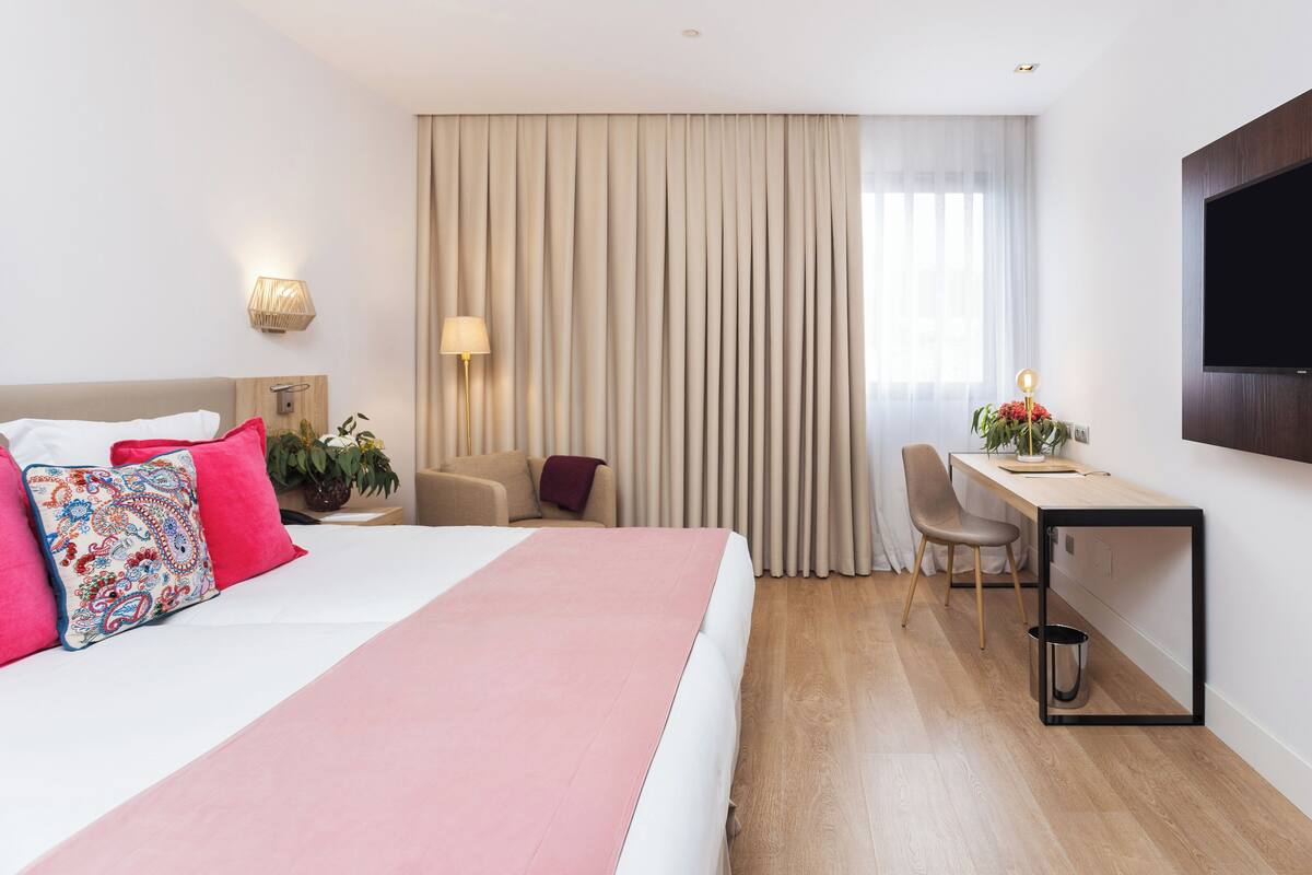 A hotel room with a bed, a desk, a chair, a TV, and a window with curtains.