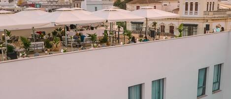 A rooftop terrace with outdoor seating, a view of the cityscape, and a building with a distinctive architectural style in the background.