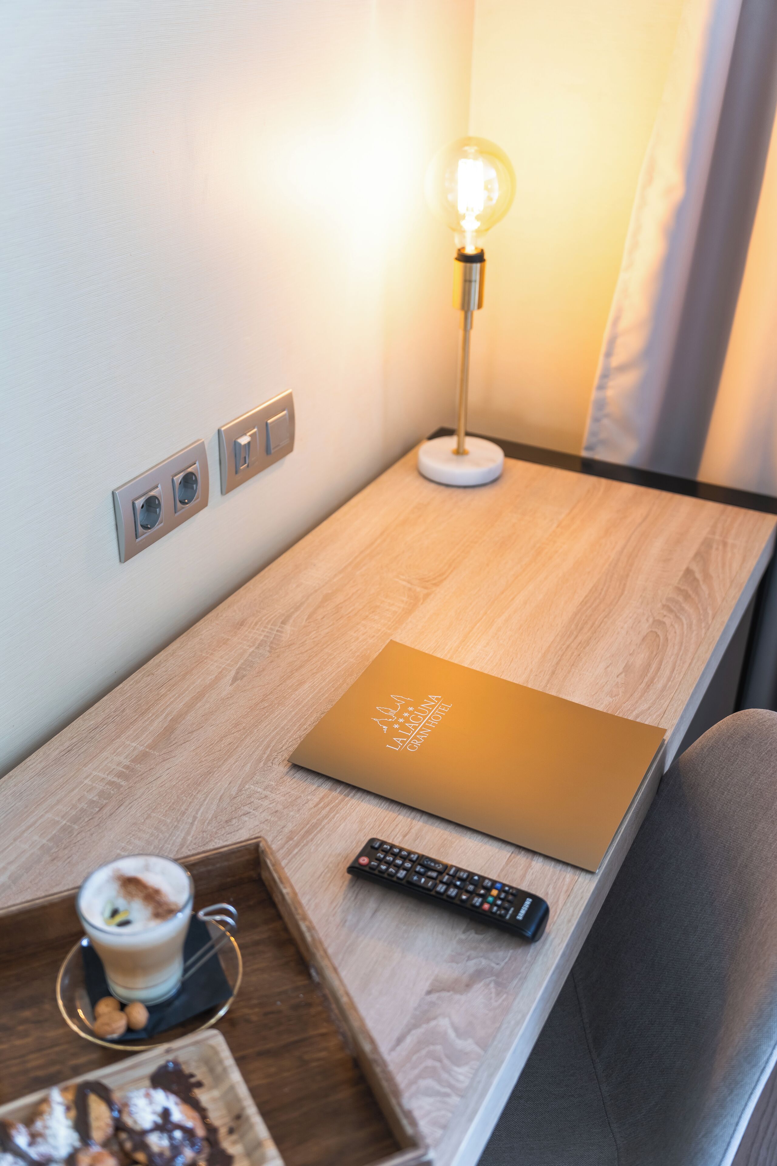 A wooden desk with a laptop, a remote control, and a tray with a drink and cookies.