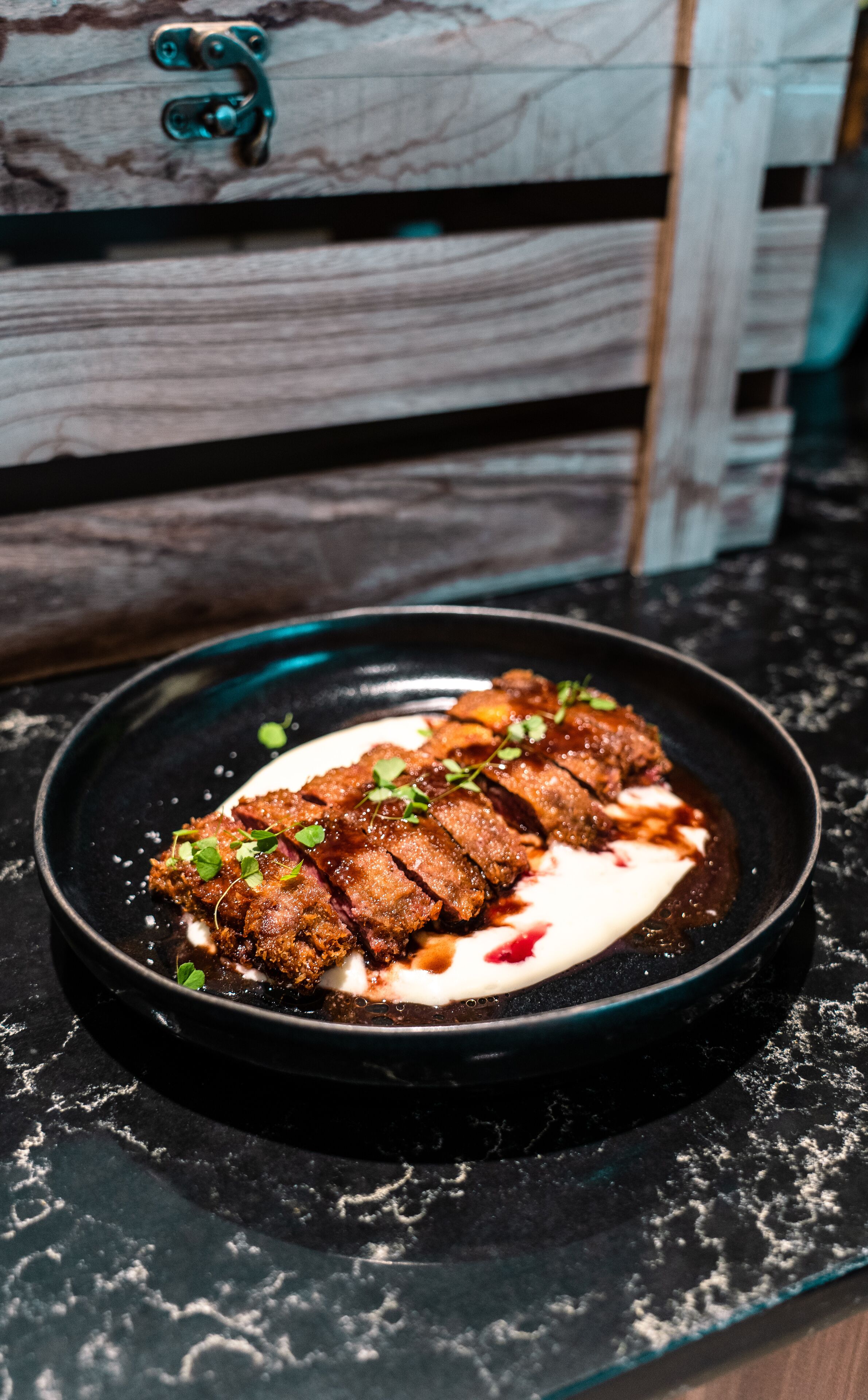 A plate of food with a dark sauce and garnish on a black plate, placed on a marble countertop.