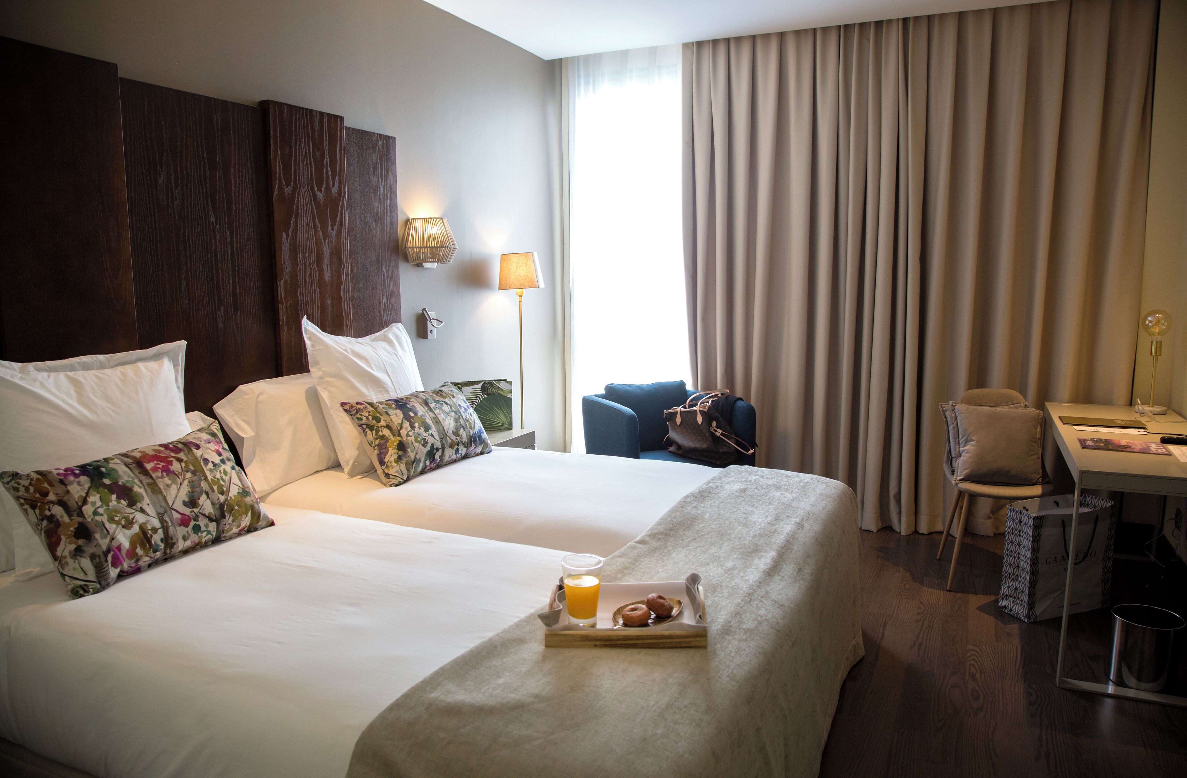 A hotel room with two beds, a desk, a chair, and a tray with a drink and pastries.