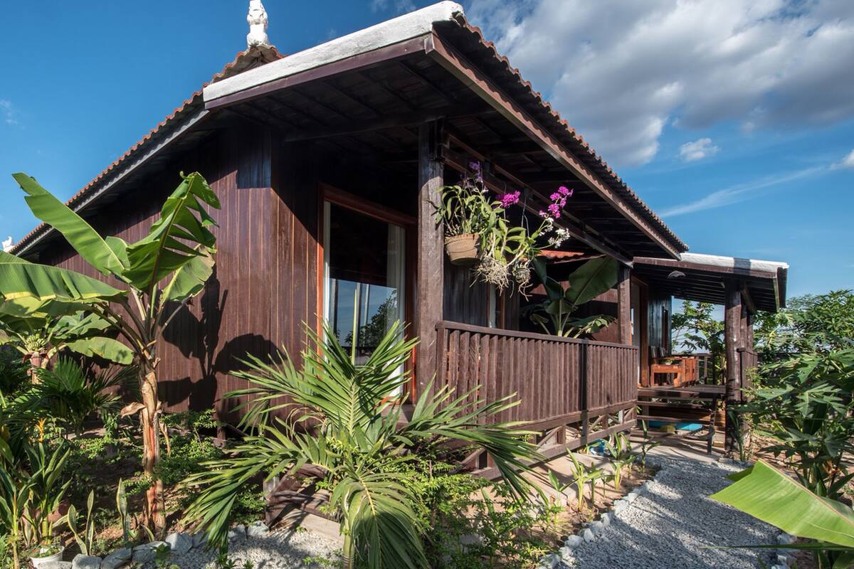 A wooden house with a porch, surrounded by tropical plants and a clear sky.