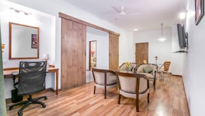 A modern living room with a wooden floor, a sofa, a dining table with chairs, a desk with a chair, a large mirror, and a ceiling fan. - Octave Hotel Double Rd (Bengaluru)