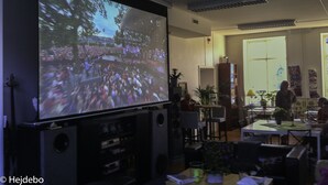 Une pièce dotée d’un grand écran diffusant un concert, d’un espace repas avec des tables et des chaises, et d’un coin cuisine équipé d’appareils électroménagers et d’un réfrigérateur.