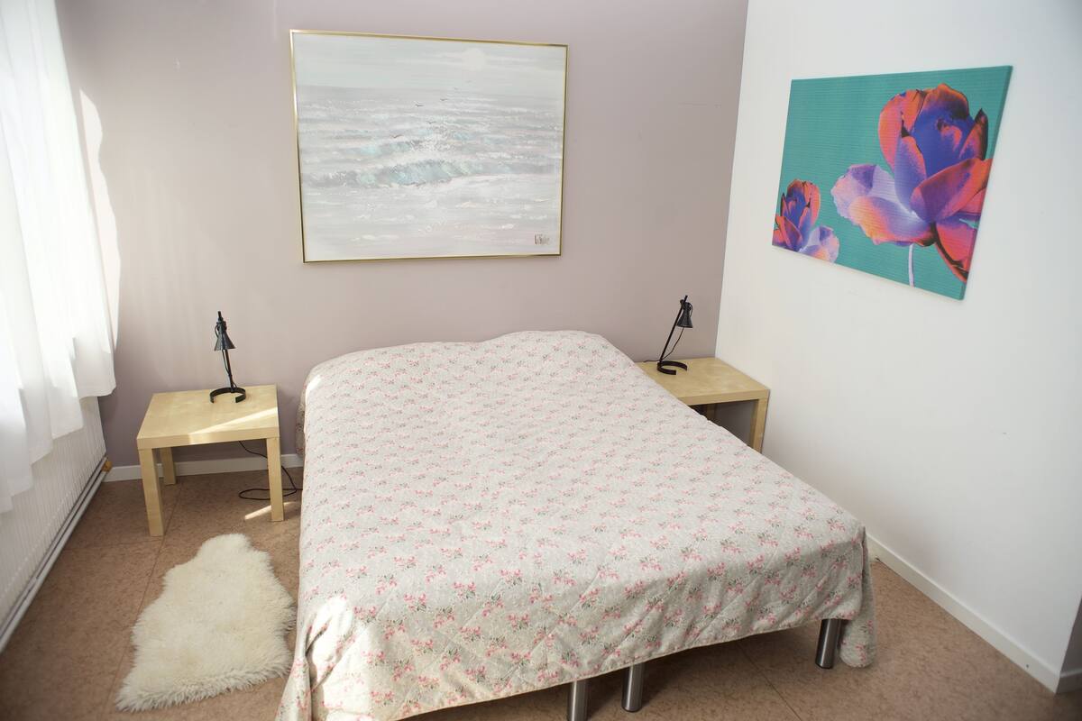 A bedroom with a bed, two bedside tables, a rug, a painting of flowers, and a painting of the sea.