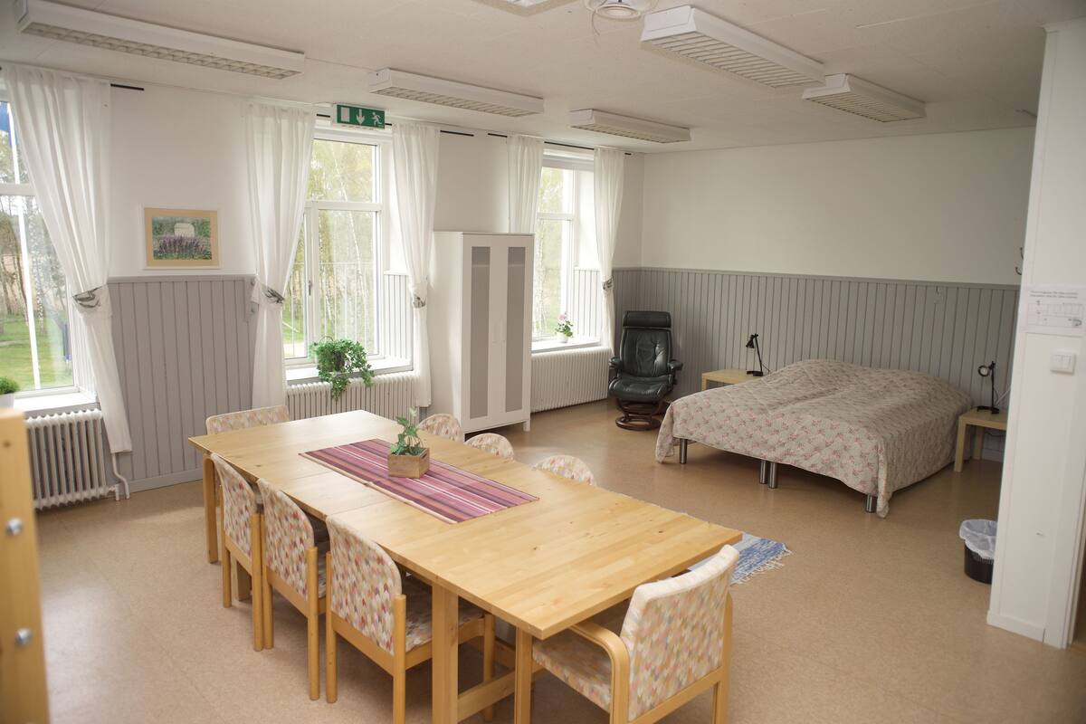A room with a wooden dining table, chairs, a bed, a radiator, and a window with curtains.