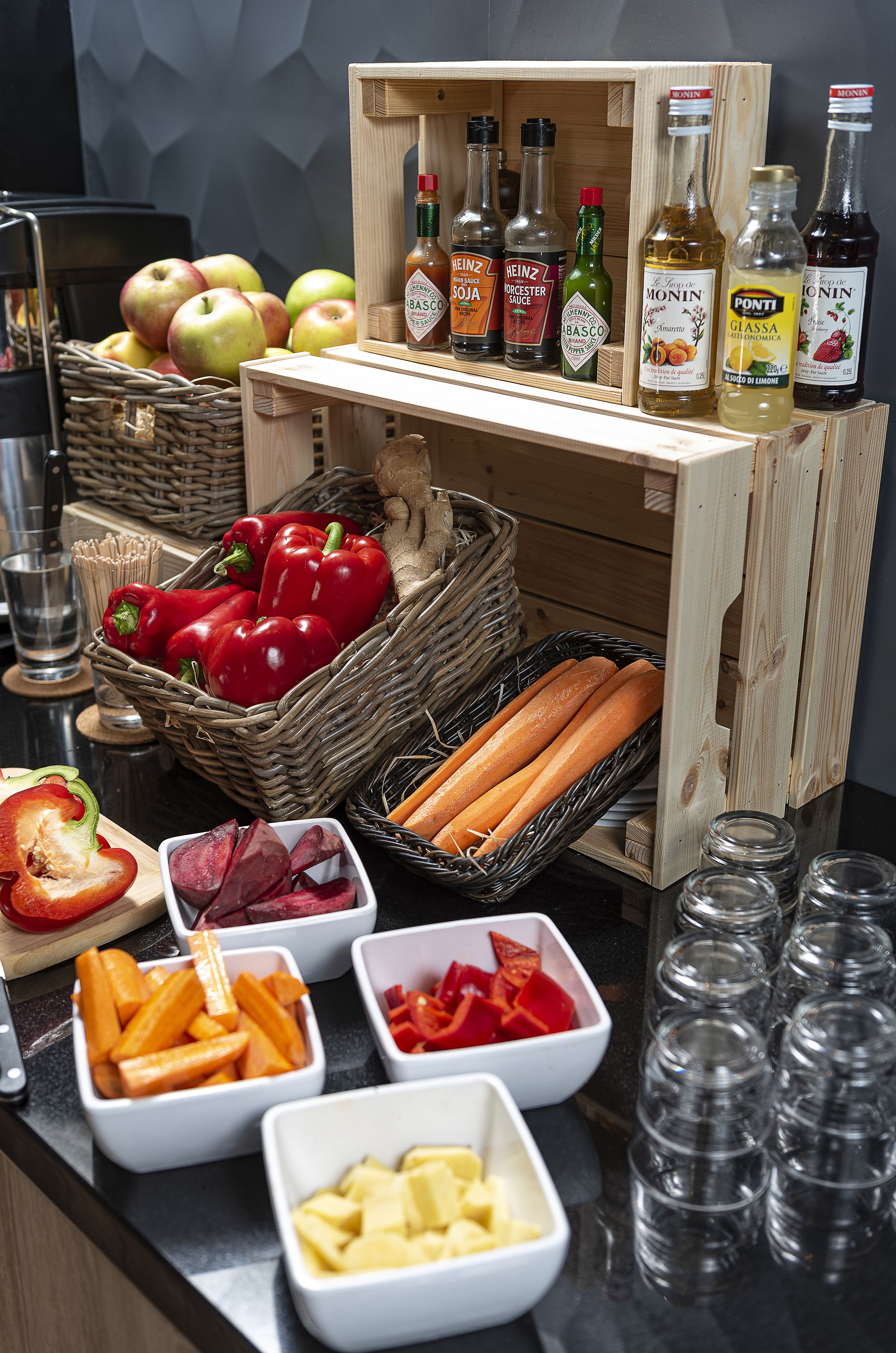 A kitchen counter with various food items and condiments, including apples, red bell peppers, carrots, and bottles of sauces.