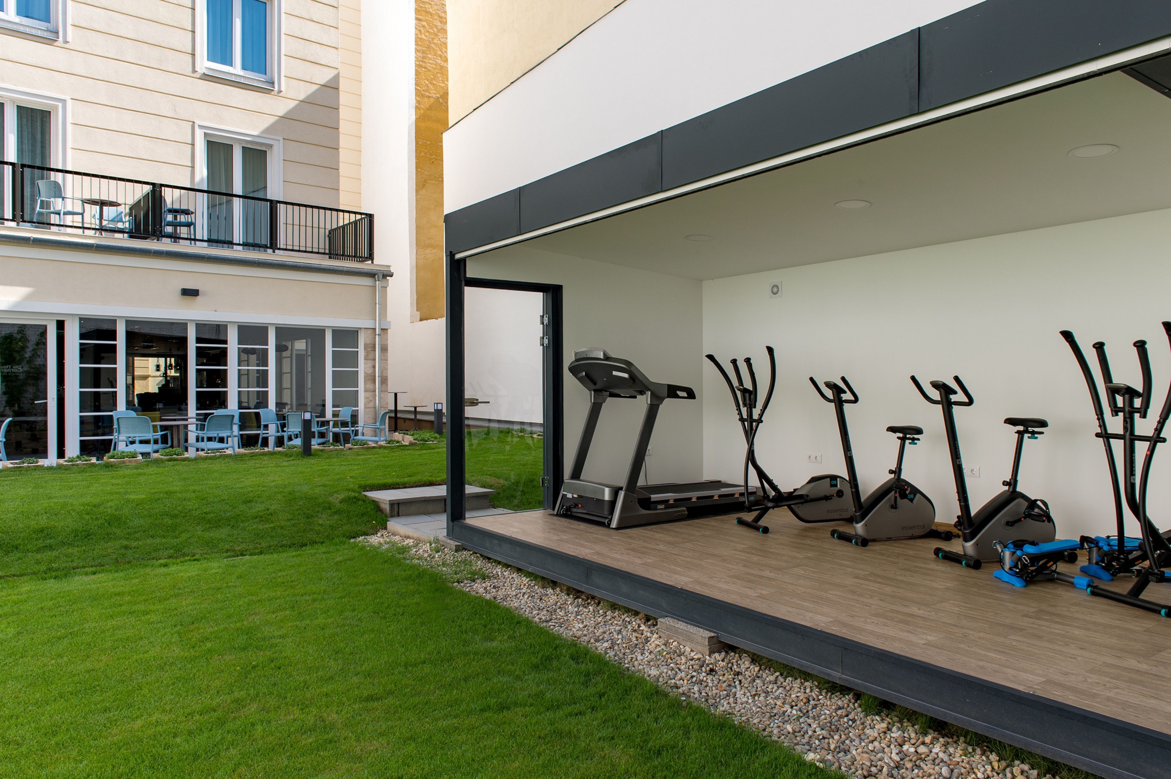 A covered outdoor area with exercise equipment, a patio with chairs, and a building with a balcony.