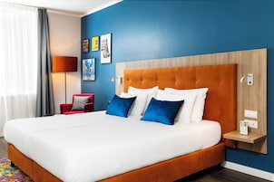 A hotel room with a large bed, a red chair, a nightstand, and artwork on the wall.