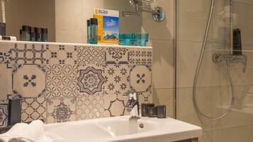 Un baño con lavamanos, espejo y ducha. Hay toallas y artículos de aseo en la encimera.