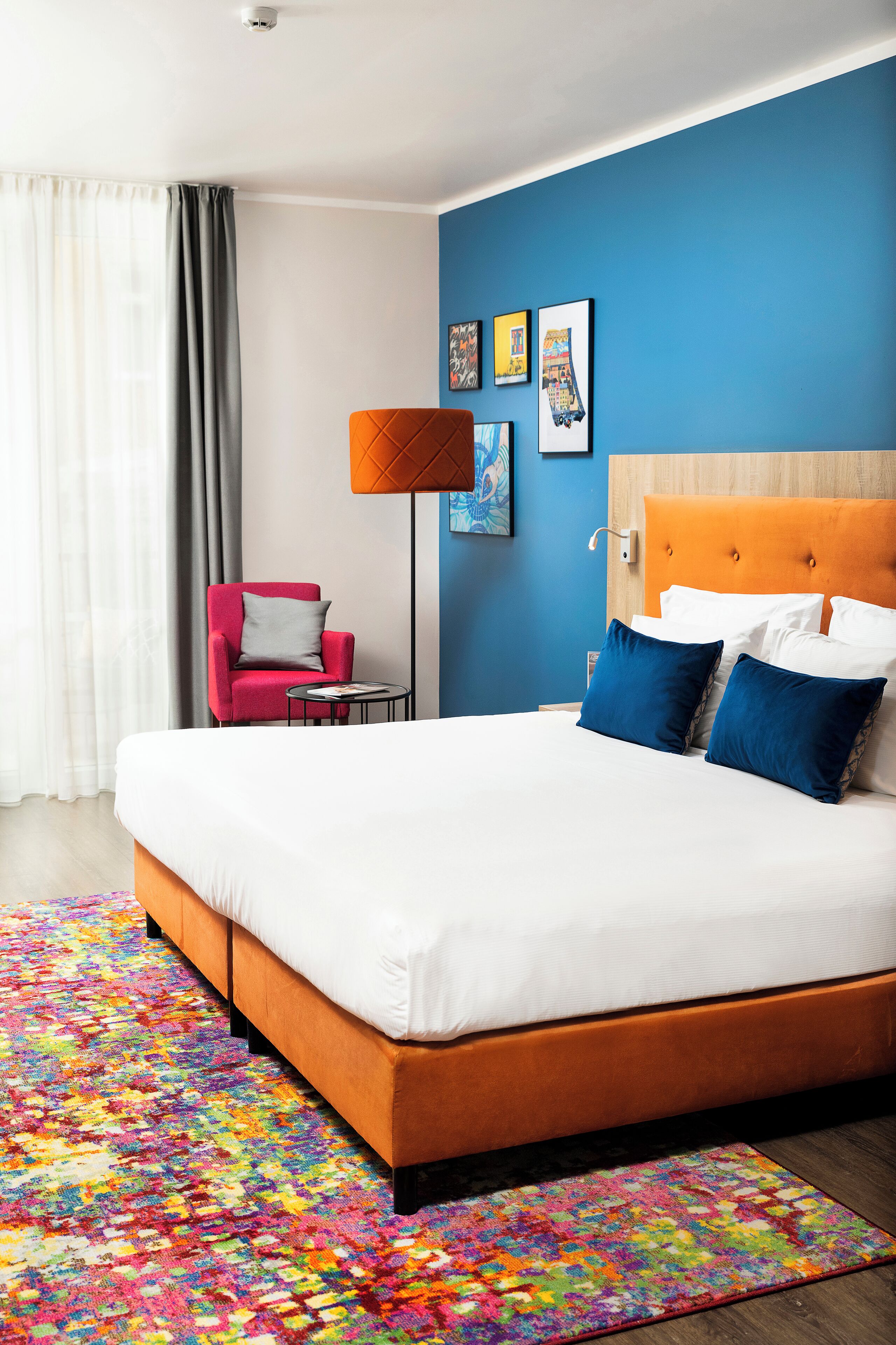 Una habitación de hotel moderna con una cama grande, un tapete colorido, un sillón rojo y una pared azul.