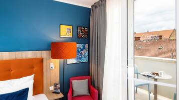 Ein Hotelzimmer mit einem Bett, einem roten Sessel, einem kleinen Tisch mit einem Buch und einer Tasse sowie einem Balkon mit Tisch und Stühlen.