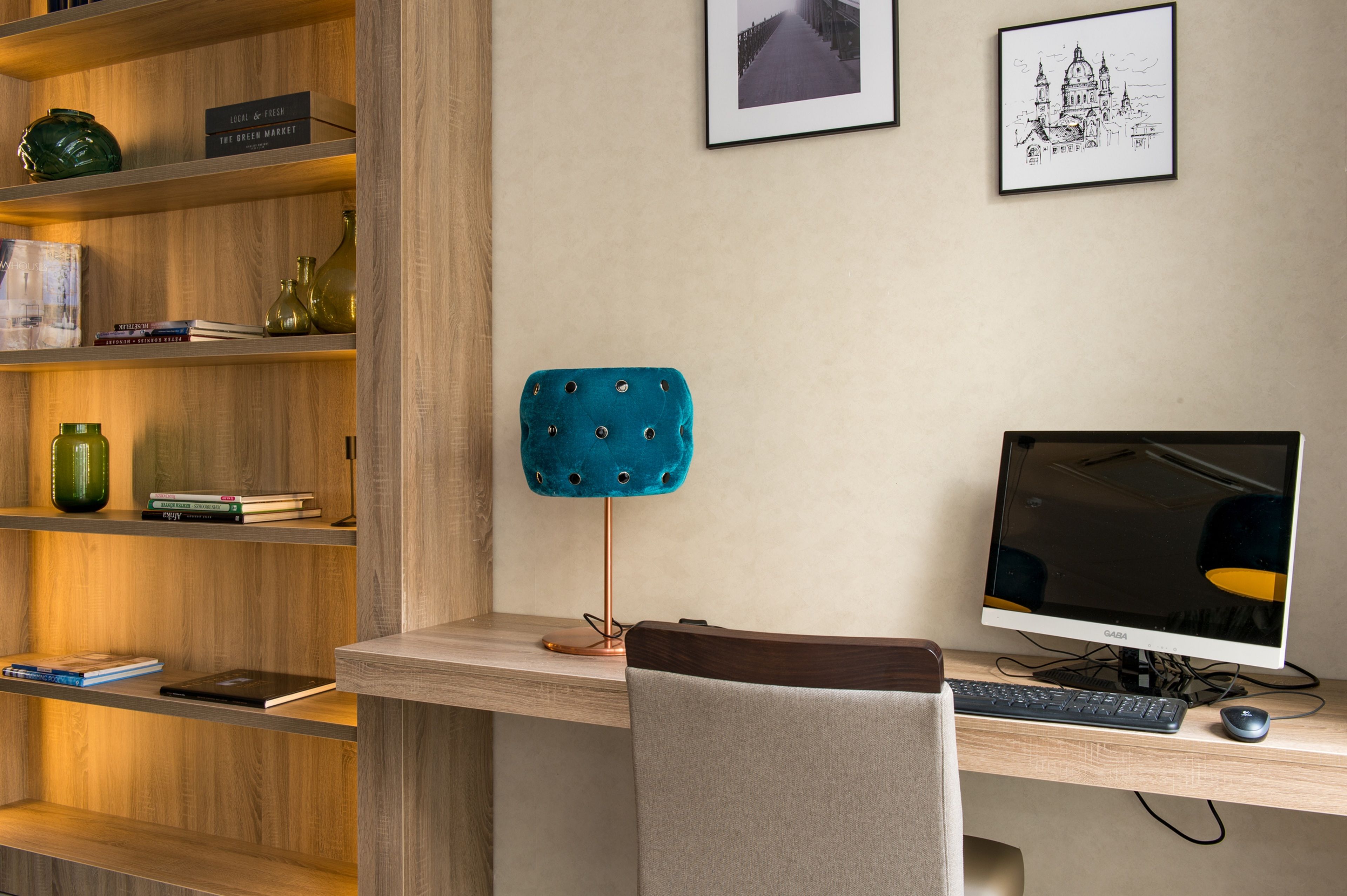 A modern workspace with a wooden desk, a teal cushioned chair, a computer monitor, keyboard, and mouse. Shelves with books and decorative items are visible.