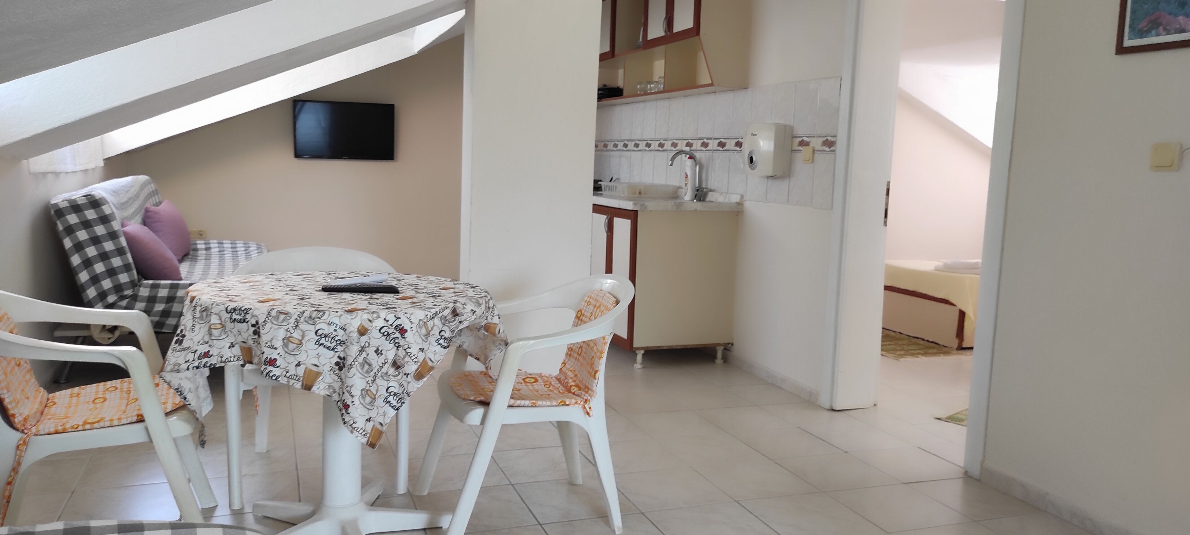 A dining area with a table and chairs, a kitchen in the background, and a television mounted on the wall.
