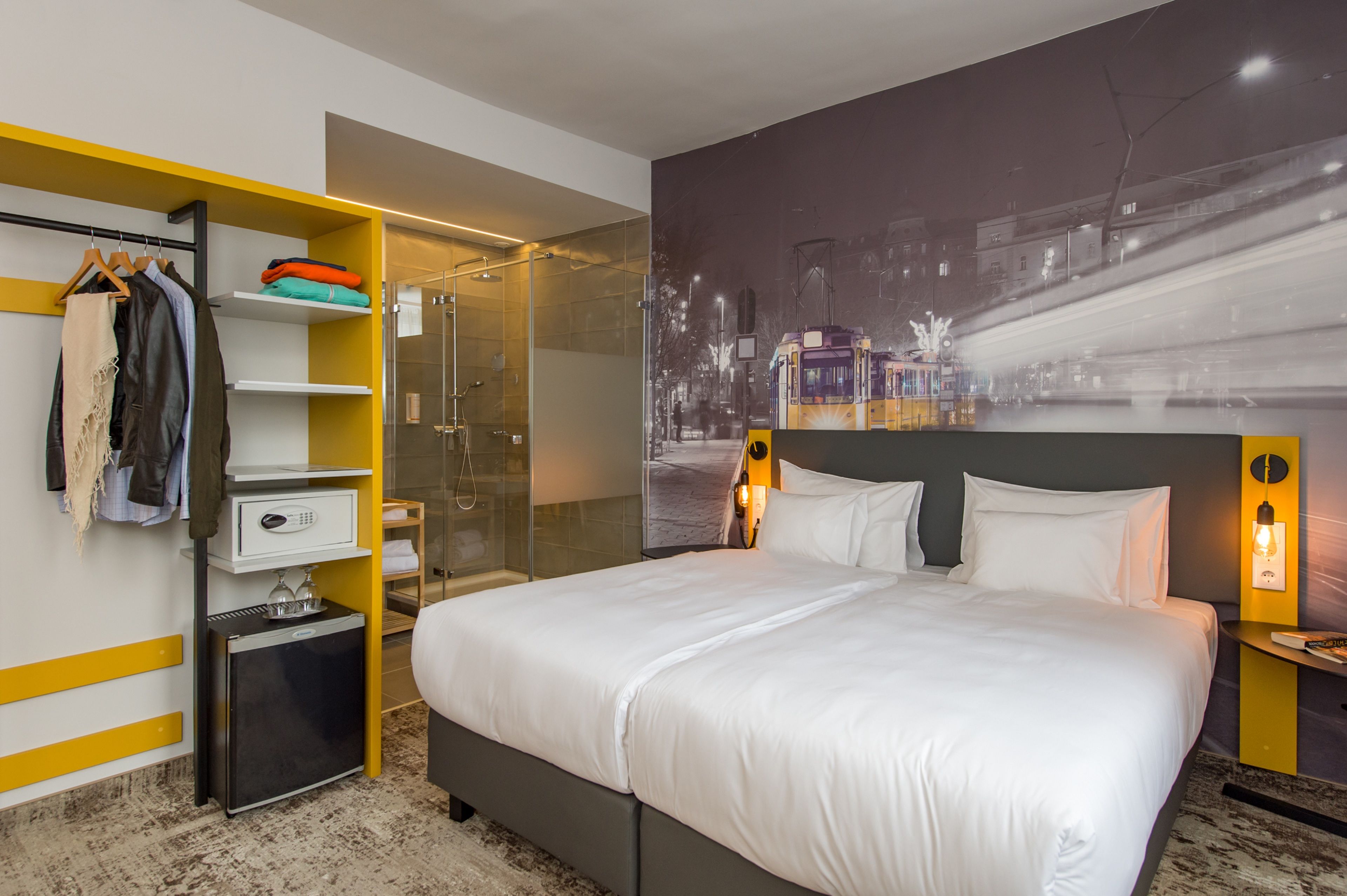 A modern hotel room with a large bed, a walk-in closet, a glass shower, and a cityscape mural on the wall.
