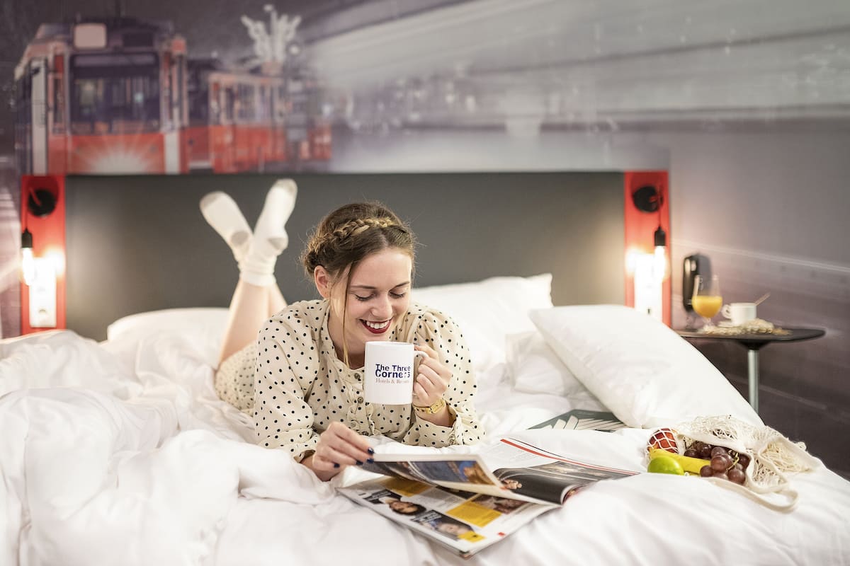 Una persona acostada en la cama leyendo una revista, con una taza, frutas y una bolsa de aperitivos sobre la mesita de noche.