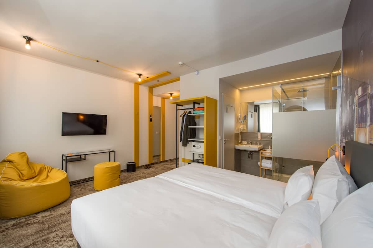 Una habitación de hotel moderna con una cama grande, un sillón de peluche amarillo, un televisor, un escritorio y un baño con espejo y lavabo.