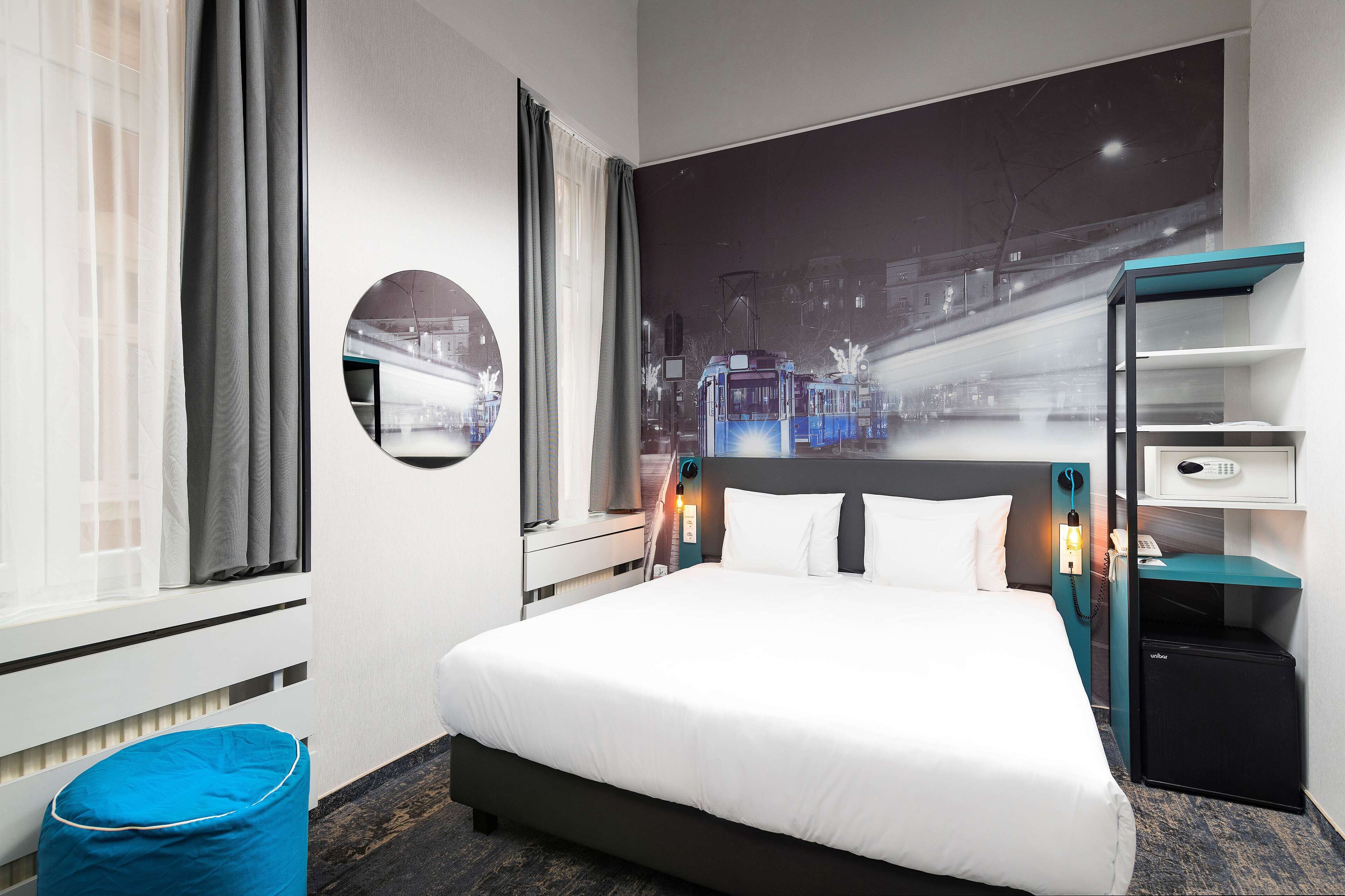 Habitación de hotel con una cama grande, un rincón de descanso, una mesita de noche con lámpara, un televisor montado en la pared y un mural con vista a la ciudad.