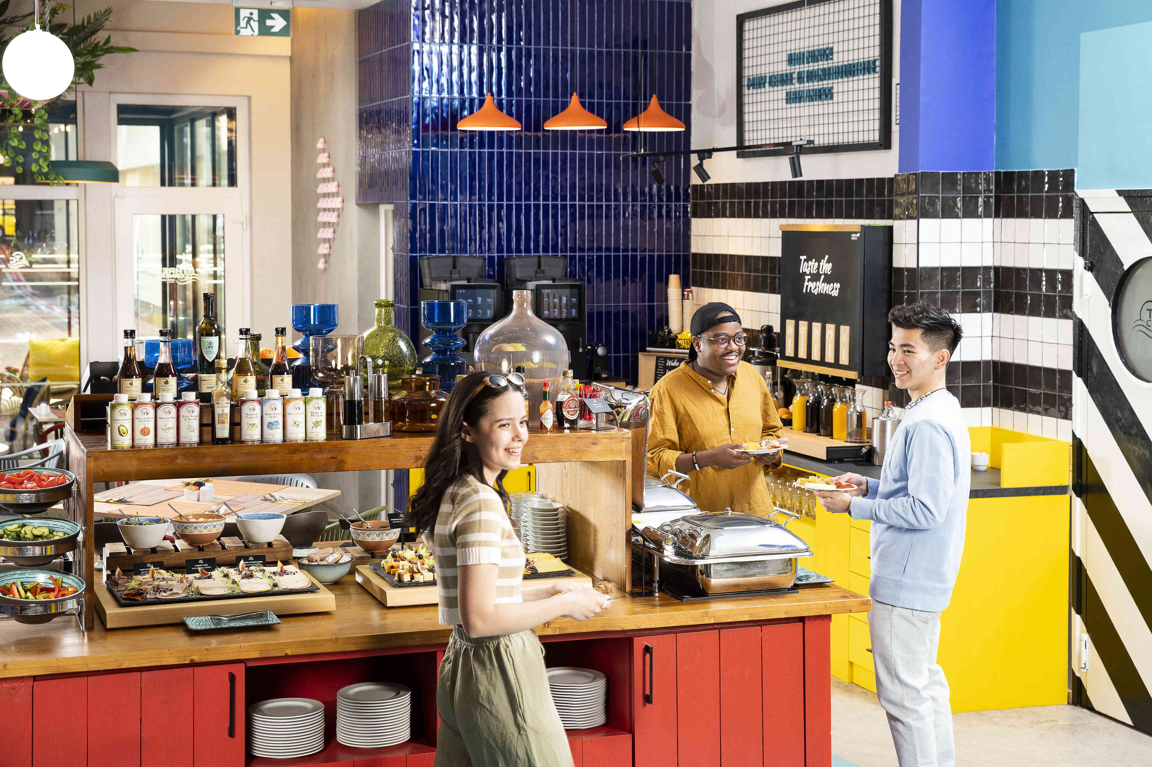 Montaje tipo bufé con diversos alimentos y bebidas, una barra atendida por personal y un área de cocina moderna al fondo.