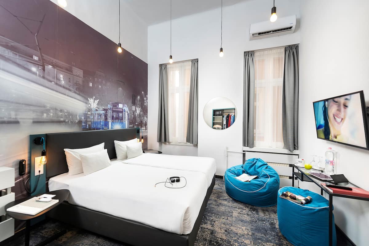 Una habitación de hotel moderna con una cama grande, un escritorio con una computadora portátil, un sillón de peluche azul y un televisor en la pared.
