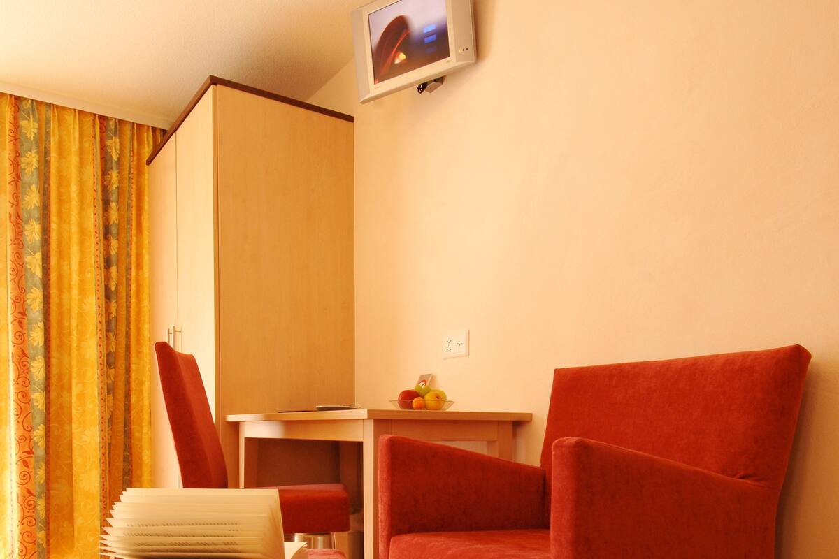 A room with two red armchairs, a small table with a fruit bowl, a TV on the wall, and a curtain with a floral pattern.